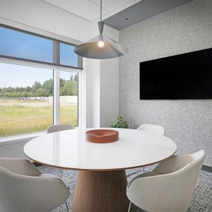 A modern meeting room with a round white table, four upholstered chairs, a wall-mounted TV, large windows overlooking a field, and potted plants in the corner.