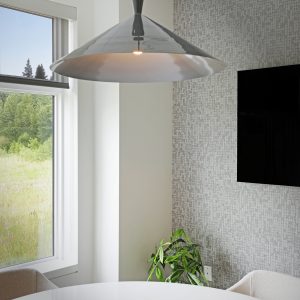 A modern dining area with a round white table, a wooden centerpiece, beige chairs, a hanging metallic pendant light, a potted plant, a wall-mounted TV, and a large window with a view of greenery outside.