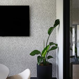 A modern office corner with a black flat-screen TV on a patterned wall, a white textured chair, a round table, and a tall leafy plant in a black planter on a gray carpet.