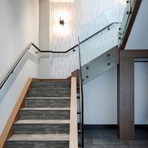 A modern indoor staircase with wooden handrails, gray carpeted steps, and glass balcony panels. The surrounding walls are light-colored with geometric patterns, and a wall-mounted light fixture is visible.