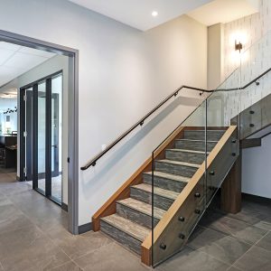 A modern indoor staircase with glass and wood railings, dark steps, and tile flooring. To the left, a glass door leads to a dining area with tables and pendant lights. Walls are light-colored and minimal decor is visible.