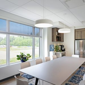 A modern office kitchen and dining area with a long table, white chairs, large windows, potted plants, pendant lights, a stainless steel fridge, and cabinets. Natural light fills the bright, clean space.