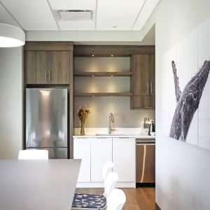 Modern kitchen with wood and white cabinets, stainless steel fridge, and sink. Open shelves hold decorative vases. Four white chairs with patterned cushions line a table. Whale artwork hangs on a pale wall. Warm, natural lighting.