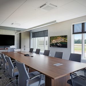 A modern conference room with a long wooden table, gray office chairs, a large wall-mounted screen, cabinets, and large windows showing an outdoor view with trees and grass.