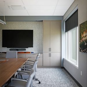 Modern conference room with a long wooden table, gray chairs, a large wall-mounted TV, cabinets, a window with a black shade, and a photo of a forested area on the wall. Neutral color palette with carpeted floor.