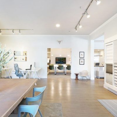 A bright, modern office space with light wood floors, white walls, a long table with blue chairs, computer desks, and display panels on the right wall. Large windows and overhead lights provide ample illumination.