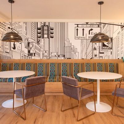 Modern café seating area with patterned bench, round white tables, cushioned chairs, teal vases, wood floor, and a black-and-white cityscape mural on the back wall. Two pendant lights hang above the tables.