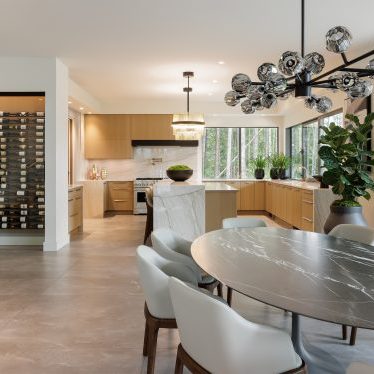 Modern dining area with a round black marble table, white cushioned chairs, a glass chandelier, wine storage wall, and open kitchen with wood cabinets. Large windows overlook trees and a patio.