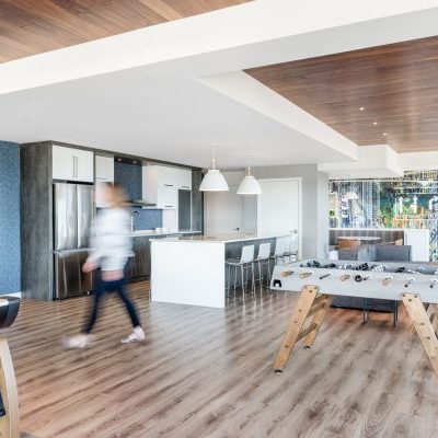 Modern open-concept living space with a kitchen, foosball table, and shuffleboard. A person walks past the kitchen island; large windows let in natural light, and a brown sofa sits in the background.