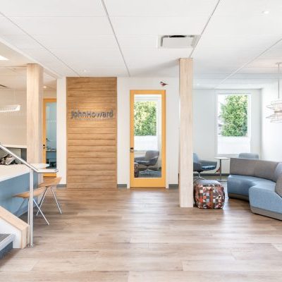 A modern, bright office lobby with wooden floors, light walls, blue and gray seating, a staircase, and a door with a "John Howard" sign. Large windows let in natural light and greenery is visible outside.