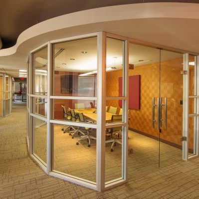 A modern office hallway with carpeted floors leads to a glass-walled conference room containing a long table and several chairs, with wood-paneled and acoustic walls inside.