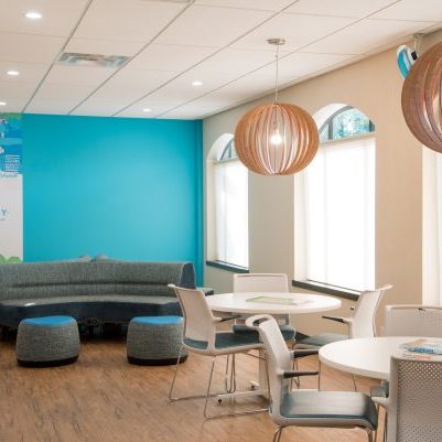 A modern, brightly lit lounge with round tables, white chairs, a curved blue sofa, two round wooden pendant lights, and large windows letting in natural light. A blue accent wall features Foundry branding and graphics.