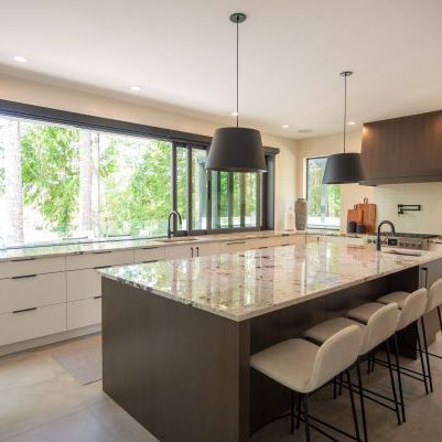 Modern kitchen with large windows, white cabinets, a marble island with four beige barstools, black pendant lights, and green trees visible outside. Countertops are clear except for some plants and kitchenware.
