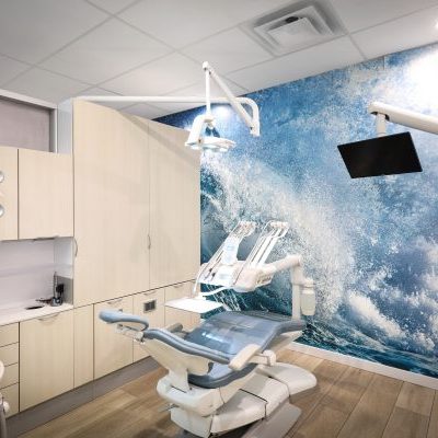 A modern dental exam room with light wood cabinets, a reclining dental chair, overhead dental lights, and a large wall mural featuring crashing ocean waves. A TV screen is mounted near the ceiling.