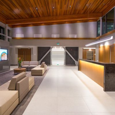 Modern, spacious lobby with beige sofas, a large dark reception desk, wood-paneled ceiling and walls, a digital display, and decorative plants; bright lighting and large windows create an inviting atmosphere.