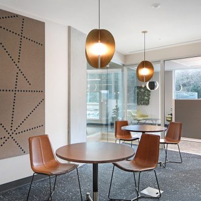 Modern office lounge with round tables, brown chairs, pendant lights, wall art with dotted lines, large windows, and a potted plant in the corner. The space is bright and inviting with neutral colors.
