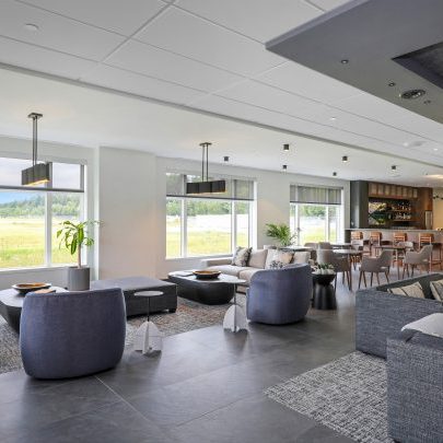 Spacious modern lounge with large windows overlooking a grassy field, featuring contemporary sofas, armchairs, coffee tables, and a bar area in the background with high stools and warm lighting.
